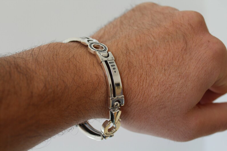 May include: A silver bracelet with a unique design is shown. The bracelet features a series of rectangular and circular links, with a contrasting black inlay. The clasp is gold-toned. The bracelet is approximately 8 inches (20 cm) in circumference.