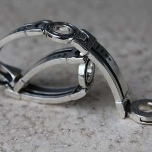 May include: A silver-toned bracelet with an intricate, geometric design. The bracelet features multiple curved bands and circular accents. The metal has a polished finish, reflecting light. The overall aesthetic is modern and industrial.