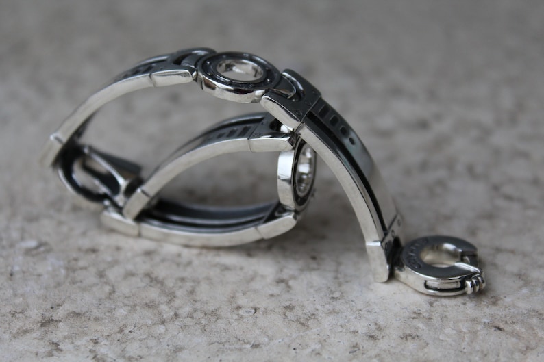 May include: A silver-toned bracelet with an intricate, geometric design. The bracelet features multiple curved bands and circular accents. The metal has a polished finish, reflecting light. The overall aesthetic is modern and industrial.