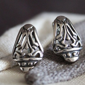 May include: A pair of silver-colored stud earrings. Each earring has a stylized, openwork design with a rounded base and a decorative pattern. The earrings are displayed on a gray and white fabric.