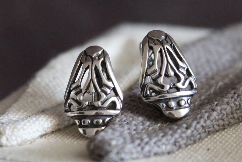 May include: A pair of silver-colored stud earrings. Each earring has a stylized, openwork design with a rounded base and a decorative pattern. The earrings are displayed on a gray and white fabric.