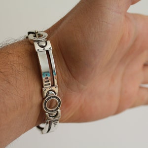 May include: A silver bracelet with a mechanical design. The bracelet features rectangular and circular elements, with the word "ALBANO" engraved on the clasp. The bracelet is shown on a wrist.