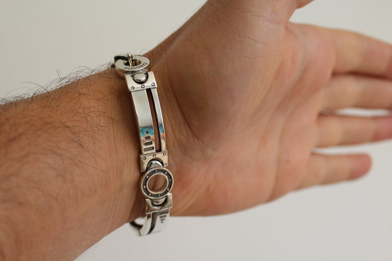May include: A silver bracelet with a mechanical design. The bracelet features rectangular and circular elements, with the word "ALBANO" engraved on the clasp. The bracelet is shown on a wrist.