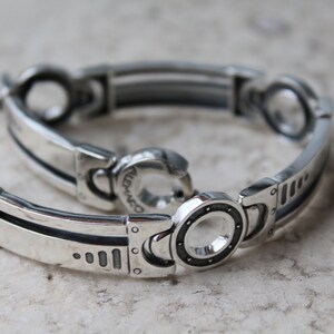 May include: A silver bracelet with a mechanical design. The bracelet features rectangular and circular links with dark accents. The clasp is a hook and eye style. The bracelet is displayed on a light-colored surface.