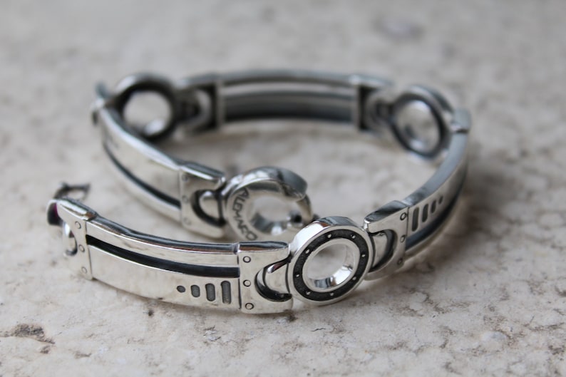 May include: A silver bracelet with a mechanical design. The bracelet features rectangular and circular links with dark accents. The clasp is a hook and eye style. The bracelet is displayed on a light-colored surface.