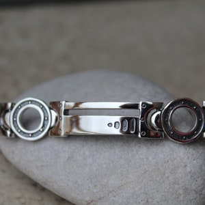 May include: A silver bracelet with a unique design. The bracelet features circular accents with a detailed pattern, connected by rectangular links. The central portion is a polished, rectangular bar. The bracelet is displayed on a gray stone.