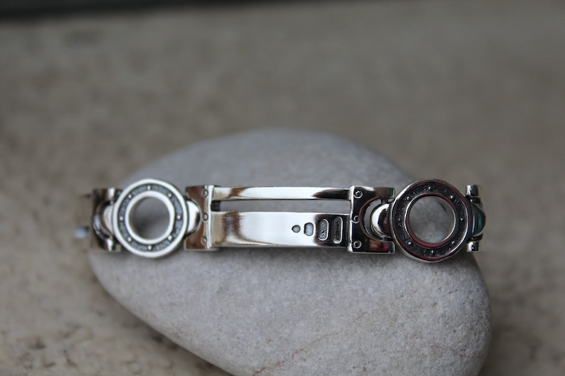 May include: A silver bracelet with a unique design. The bracelet features circular accents with a detailed pattern, connected by rectangular links. The central portion is a polished, rectangular bar. The bracelet is displayed on a gray stone.