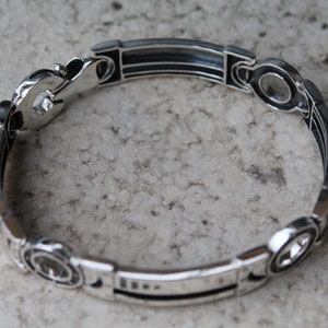 May include: A silver bracelet with an intricate design. The bracelet features a series of rectangular and circular links connected by decorative elements. The metal has a polished finish, reflecting light. The bracelet is shown on a neutral background.