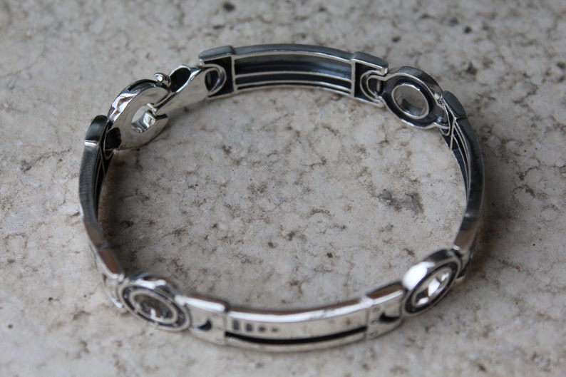 May include: A silver bracelet with an intricate design. The bracelet features a series of rectangular and circular links connected by decorative elements. The metal has a polished finish, reflecting light. The bracelet is shown on a neutral background.