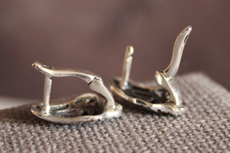 May include: Close-up of two silver-colored earrings with a lever back closure. The earrings are detailed with a textured design and are set against a neutral background. The focus is on the intricate design and the clasp mechanism.