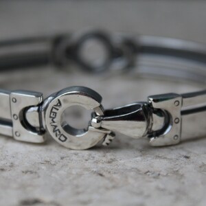 May include: A silver-toned bracelet with a unique clasp design. The bracelet features rectangular links with a grooved center and rivet details. The clasp has the word "ALEMARD" engraved on it.