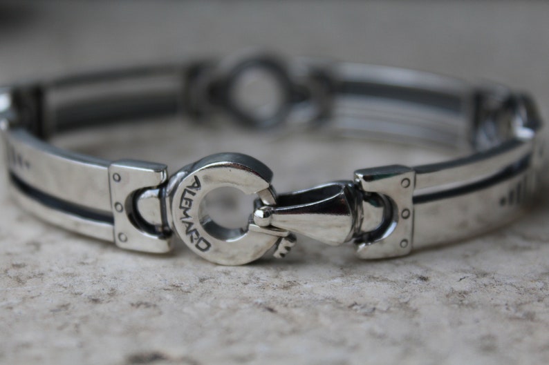 May include: A silver-toned bracelet with a unique clasp design. The bracelet features rectangular links with a grooved center and rivet details. The clasp has the word "ALEMARD" engraved on it.