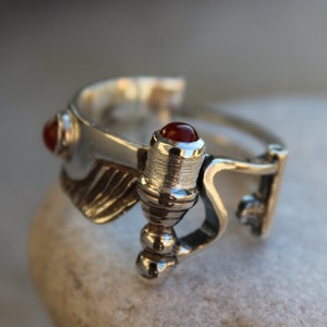 May include: A silver ring with a red gemstone and a detailed design. The ring features a wing-like shape and a small, cylindrical element with a red gemstone.