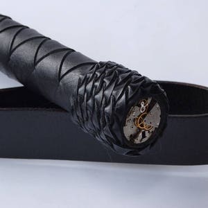 May include: A black leather whip with a braided handle and a decorative metal detail at the end. The metal detail is a circular design with gears and other intricate details.