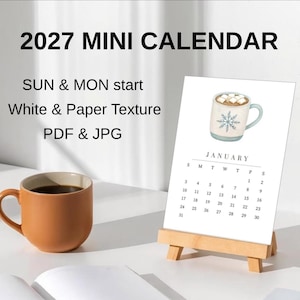 Può includere: Un mini calendario 2027 su un cavalletto in legno, con una pagina di gennaio che mostra un'illustrazione ad acquerello di una tazza piena di cioccolata calda e marshmallow. Il testo include SUN & MON start, White & Paper Texture, PDF & JPG.