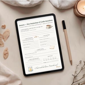 May include: A digital tablet displays a January planner with the text "Slow Beginnings & Gentle Intentions." A pen, a mug, and a lit candle are on the off-white surface. Decorative elements include dried flowers and small wooden shapes.