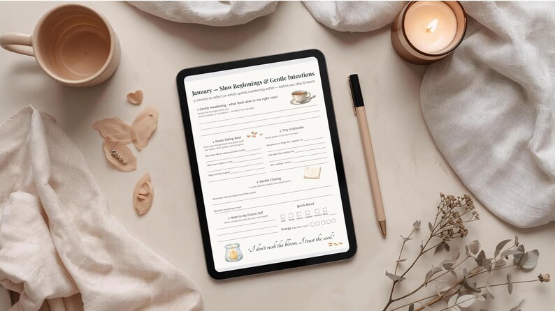 May include: A digital tablet displays a January planner with the text "Slow Beginnings & Gentle Intentions." A pen, a mug, and a lit candle are on the off-white surface. Decorative elements include dried flowers and small wooden shapes.
