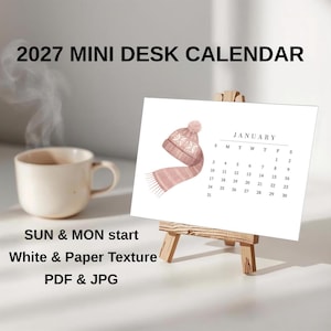 Può includere: Un mini calendario da scrivania 2027 su un piccolo cavalletto in legno. La pagina del calendario presenta un'illustrazione di un berretto e una sciarpa in maglia rosa accanto alla griglia di gennaio. Una tazza in ceramica beige è a sinistra.