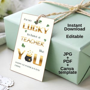Może przedstawiać: Etykieta prezentowa z napisem "I'M SO LUCKY to have a TEACHER like YOU" w złotych literach, z akcentami koniczyny. Etykieta jest przymocowana do jasnozielonego pudełka prezentowego z naturalną kokardą ze sznurka. Etykieta mówi również "Happy St Patrick's Day".