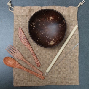 Op de afbeelding: Een set milieuvriendelijke eetartikelen, waaronder een kokosnootschaal, houten bestek, een bamboe rietje en een schoonmaakborstel, gepresenteerd op een jute zak. De houten items zijn bruin.