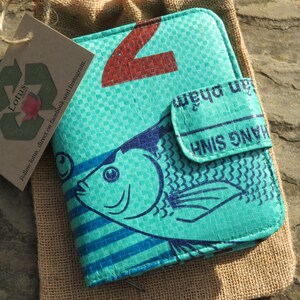 May include: A turquoise blue wallet with a fish design and the text "H&Agrave;NG SINH" and "jan phẩm" printed on it. The wallet is made from recycled materials and has a zipper closure.