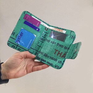 May include: A green wallet made from recycled materials. The wallet is folded closed and has several cards inside. The wallet has text in Vietnamese on the outside.