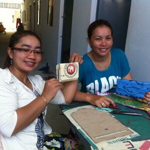 May include: Two women are working on a craft project. One woman is holding a small, brown, rectangular box with a white elephant logo and the text "Elephant Brand" printed on it. The other woman is holding a blue fabric wallet with a white fish design. Both women are sitting at a table with various craft supplies, including fabric scraps, scissors, and a cardboard template.