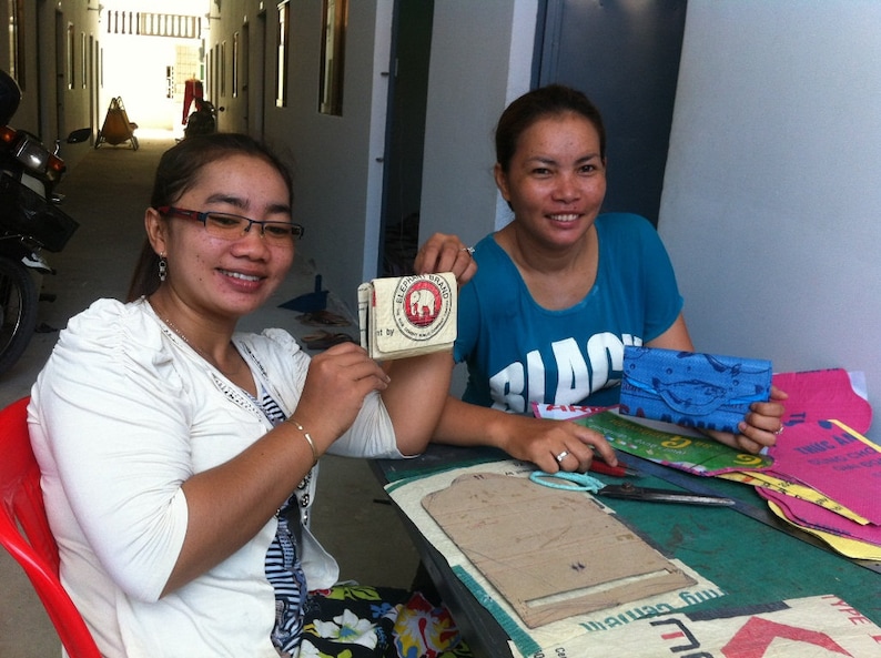 May include: Two women are working on a craft project. One woman is holding a small, brown, rectangular box with a white elephant logo and the text "Elephant Brand" printed on it. The other woman is holding a blue fabric wallet with a white fish design. Both women are sitting at a table with various craft supplies, including fabric scraps, scissors, and a cardboard template.