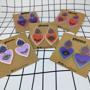 Può includere: Una collezione di orecchini a forma di cuore colorati con disegni di occhi e ciglia. Gli orecchini sono in tonalità di viola, arancione e verde, con un accento di stampa leopardata. Ogni paio è esposto su una carta marrone con il marchio "glucose".