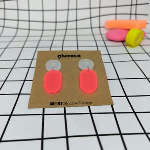 Può includere: Un paio di orecchini rosa neon e bianchi. Gli orecchini sono in acrilico e hanno una parte superiore circolare e una inferiore ovale. Sono esposti su una carta marrone con il testo "glucose" e "GlucoseDesign".
