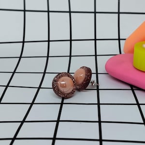 May include: A pair of stud earrings featuring a light peach-colored center surrounded by a glittery, dark brown border. The earrings have a post backing and are set against a black and white grid background.