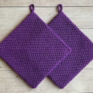 May include: Two purple crocheted pot holders with a loop handle on each. The pot holders are made with a textured stitch.