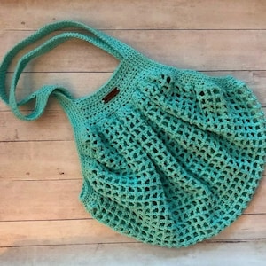 May include: A teal blue crocheted tote bag with a brown leather handle. The bag is made with a mesh pattern and is perfect for carrying groceries or other items.
