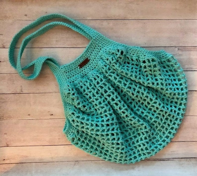 May include: A teal blue crocheted tote bag with a brown leather handle. The bag is made with a mesh pattern and is perfect for carrying groceries or other items.