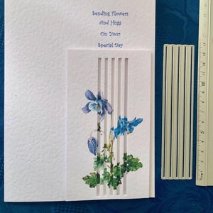 Könnte beinhalten: Eine weiße Grußkarte mit dem blauen Text "Sending Flowers And Hugs On Your Special Day". Sie zeigt ein blaues und grünes Blumenmuster und einen vertikalen Streifenausschnitt. Ein Lineal liegt neben der Karte.
