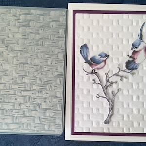 May include: A greeting card with two blue and pink birds on a branch, set against a white, textured backdrop. The card has a purple border and a clear, textured plastic cover.