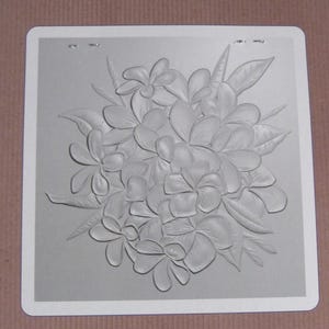 May include: A gray square card with a white border features a detailed embossed design of plumeria flowers and leaves. The floral arrangement is centered on the card, creating a three-dimensional effect. The card is set against a neutral background.