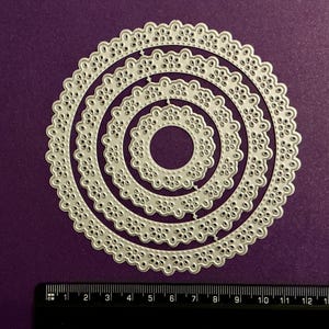 May include: A set of five nested, die-cut metal circles with floral and scalloped edges. The silver-coloured die cuts are arranged on a purple surface. A ruler with centimetres is at the bottom.
