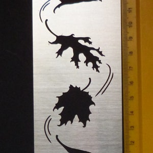 May include: A metal stencil with four black silhouettes of leaves blowing in the wind. The stencil is 17.5 cm tall and has the text "620101 ynoll Hatano" printed on the bottom.