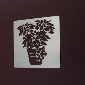 May include: A white metal stencil of a poinsettia plant in a pot with a bow. The stencil is designed for crafting and decorating.