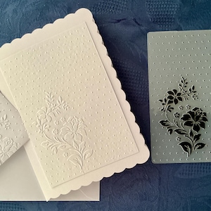 May include: A white scalloped-edge card and matching envelope featuring an embossed floral and dot pattern, displayed alongside a rectangular translucent plastic embossing folder that shows the same floral and dot design in black.