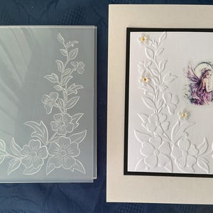 May include: A rectangular clear plastic embossing folder featuring a floral vine pattern and a finished white greeting card with a black border, embossed with the same floral design and a small sticker of a fairy with purple wings.