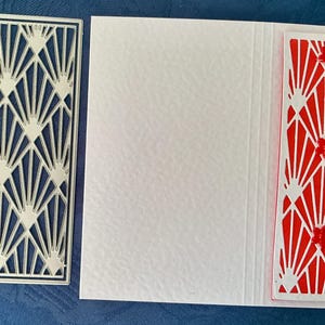 May include: A silver metal rectangular die-cut stencil featuring a geometric fan pattern, positioned next to a black ruler showing 6 inches in length. A white cardstock panel displays the same intricate fan design layered over a red background.