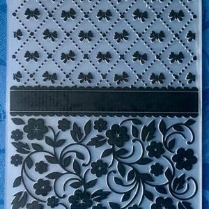 Könnte beinhalten: Eine transparente Plastikfolie mit zwei verschiedenen Designs. Das obere Design ist ein sich wiederholendes Muster aus schwarzen Schleifen in einer Diamantform. Das untere Design ist ein sich wiederholendes Muster aus schwarzen Blumen und Ranken.
