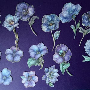 May include: A collection of watercolor-style floral cutouts in shades of blue, purple, and green. The flowers vary in shape and size, with gold accents and green leaves. They are arranged on a dark purple background.