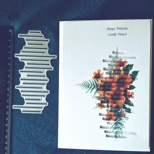 Puede incluir: Una tarjeta de cumpleaños blanca con el texto "Happy Birthday Lovely Friend" y un diseño floral. Una herramienta de corte de metal está al lado de la tarjeta. La tarjeta presenta un diseño recortado con flores naranjas y hojas verdes.