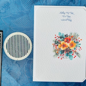 May include: A white greeting card with the text "Thinking Of You On Your Special Day" in blue script. The card features a floral design with orange, yellow, and red flowers. A metal die-cut circle is next to the card.
