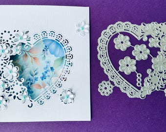 May include: A white card with a heart-shaped cutout, adorned with delicate floral embellishments and a floral patterned insert. A separate die-cut heart with floral designs sits beside it, perfect for crafting projects.