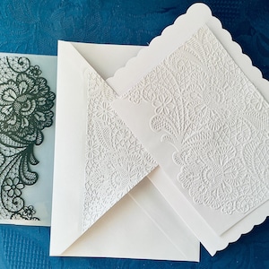 Peut inclure: Une carte rectangulaire blanche à bord festonné avec un motif de dentelle florale en relief, accompagnée d'une enveloppe blanche assortie avec un rabat intérieur en dentelle, et un pochoir en plastique translucide à motif dentelle noire.