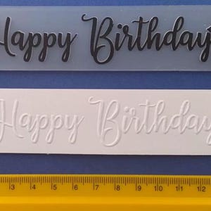 May include: Two rectangular molds with the words "Happy Birthday" in cursive script. The top mold is translucent with black text and floral accents. The bottom mold is white with embossed text and floral accents. A yellow ruler is at the bottom.